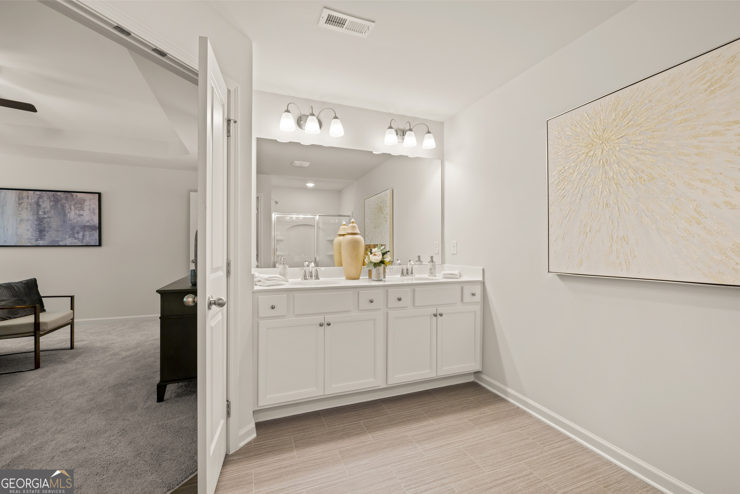 Bathroom with a white double-sink vanity, large mirror, and doorway opening to a carpeted bedroom.