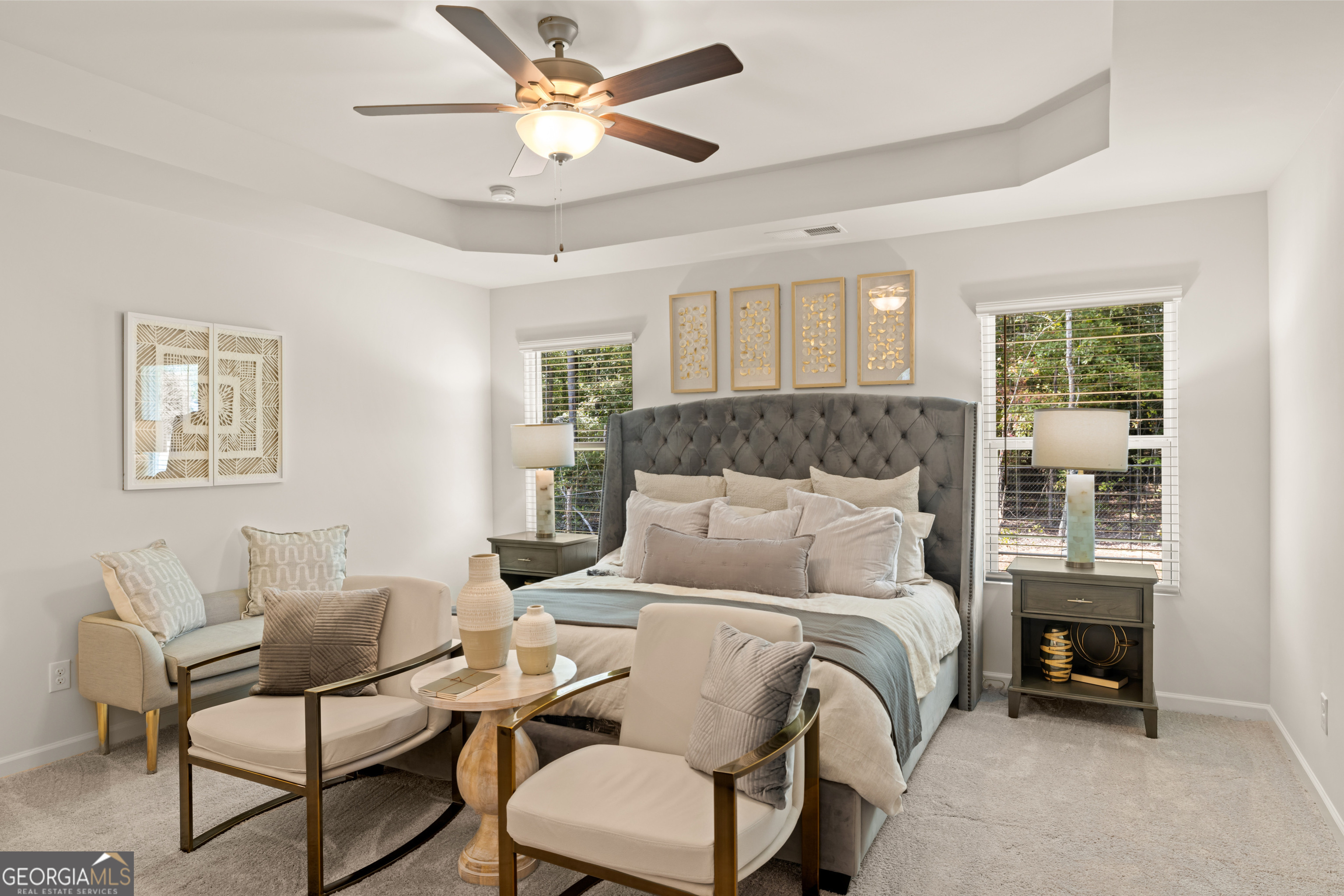Staged bedroom with a gray tufted bed, neutral seating, and a tray ceiling with a ceiling fan.