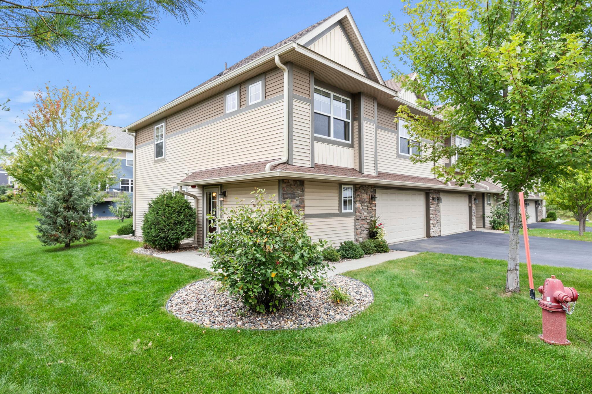 Beige two-story attached home with stone accents, white garage doors, and landscaped lawn.