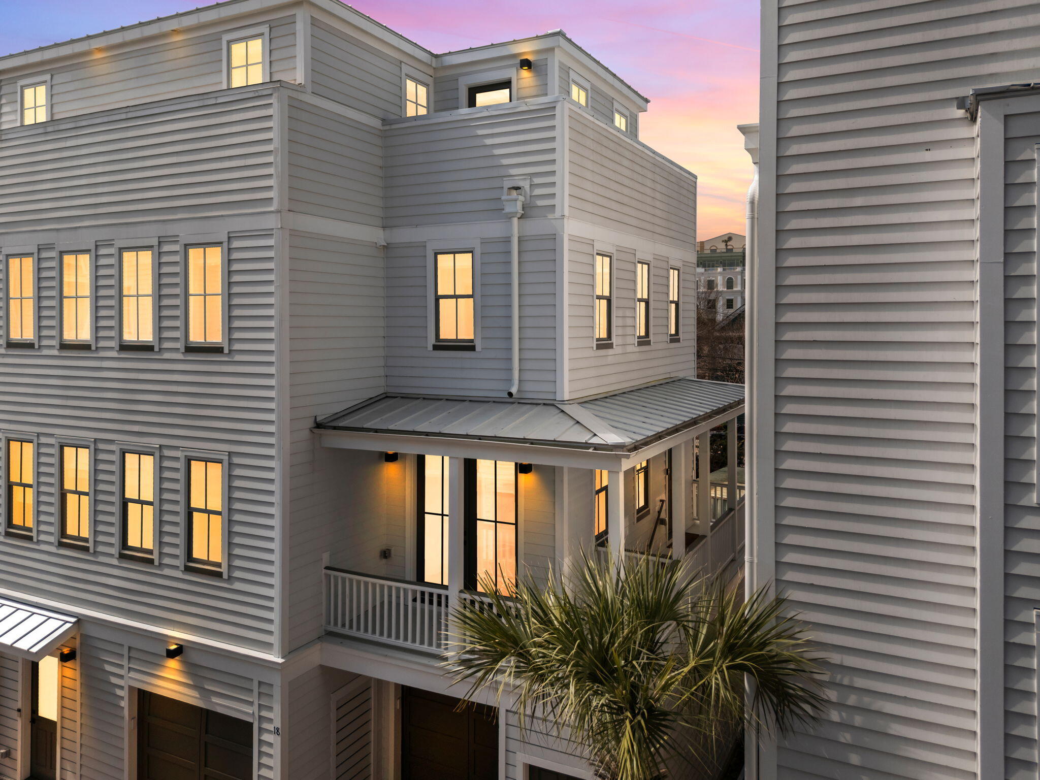 Gray multi-story home with a metal porch roof and glowing windows at dusk.