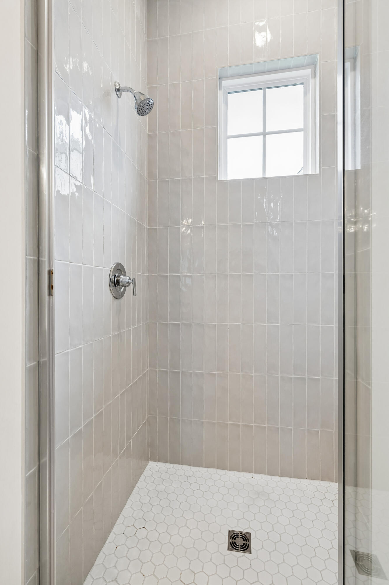 Tiled walk-in shower with glass enclosure, chrome fixtures, hexagonal floor tile, and a small window.