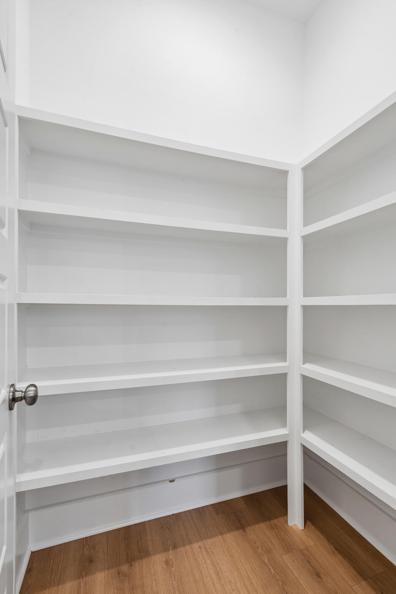 Empty pantry with white wraparound shelving and wood-look flooring.