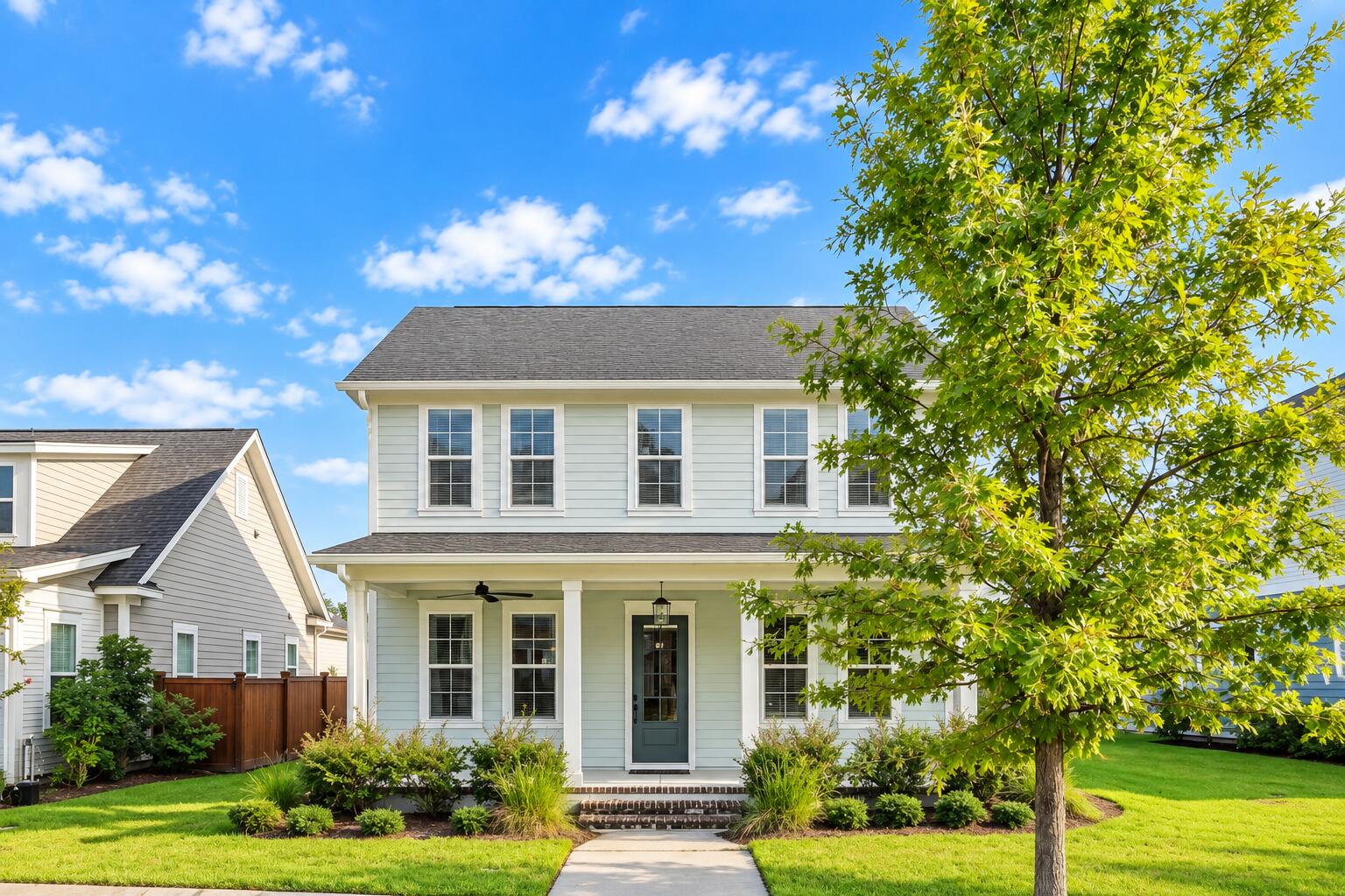 Two-story light blue house with a covered porch, landscaped lawn, and large leafy tree.