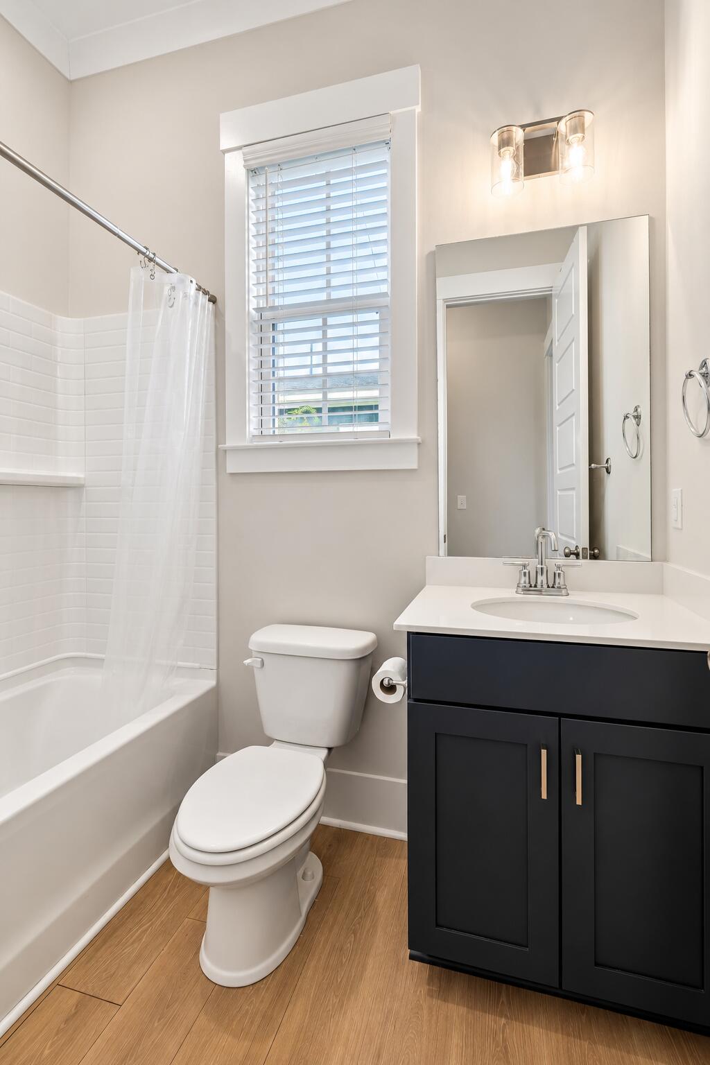 Bathroom with a white tub and shower, toilet, dark vanity, and window with blinds.