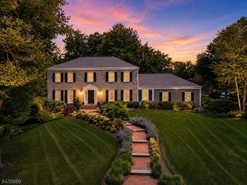 Two-story home with glowing windows, landscaped lawn, and a curved brick walkway at dusk.