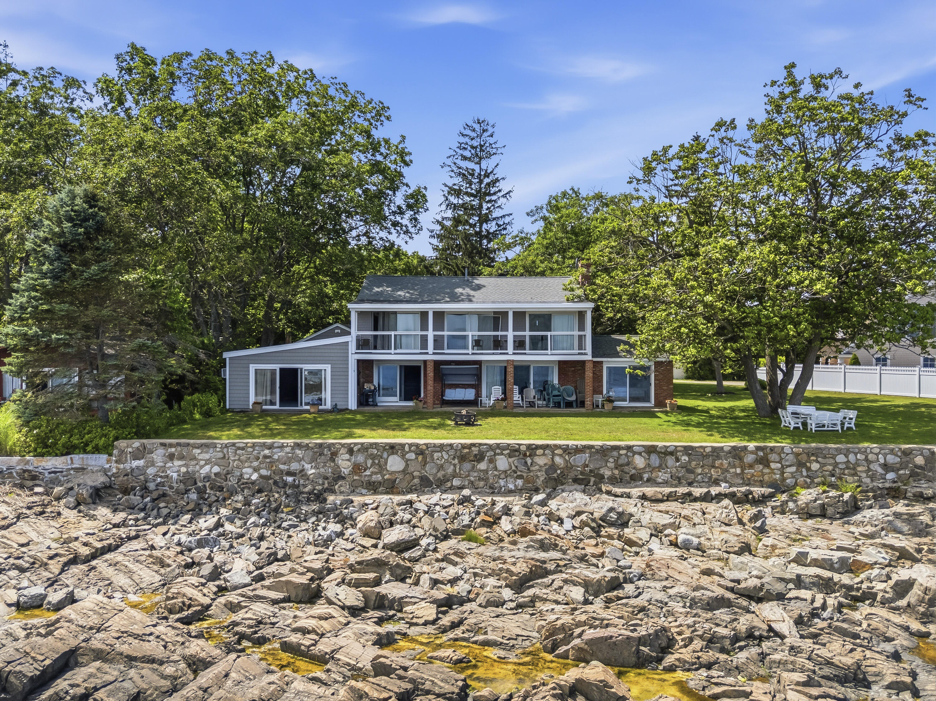 Two-story coastal house above a rocky shoreline, with a stone seawall and tree-lined lawn.