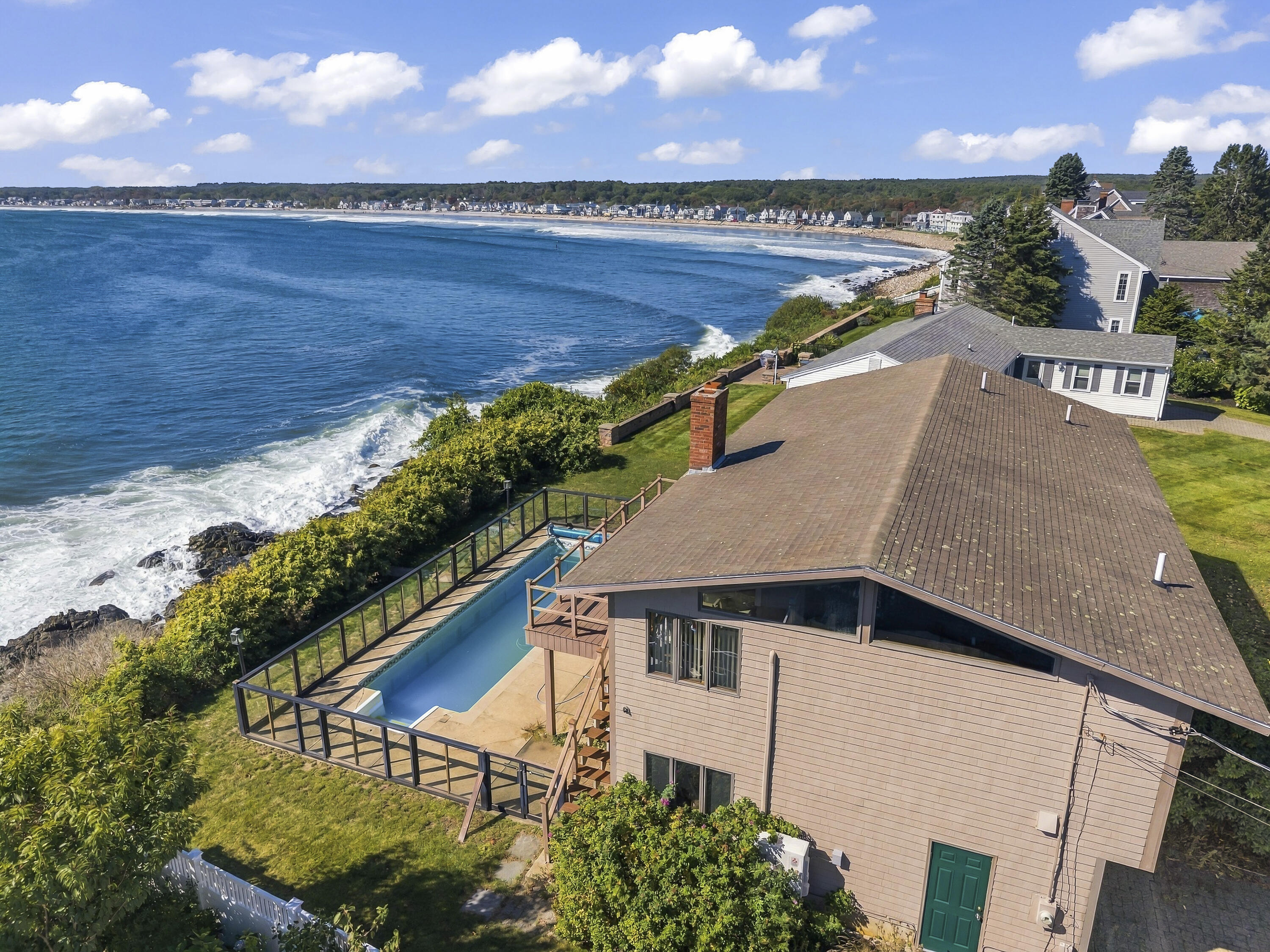 Coastal home with a fenced pool overlooking rocky oceanfront and a distant sandy beach.