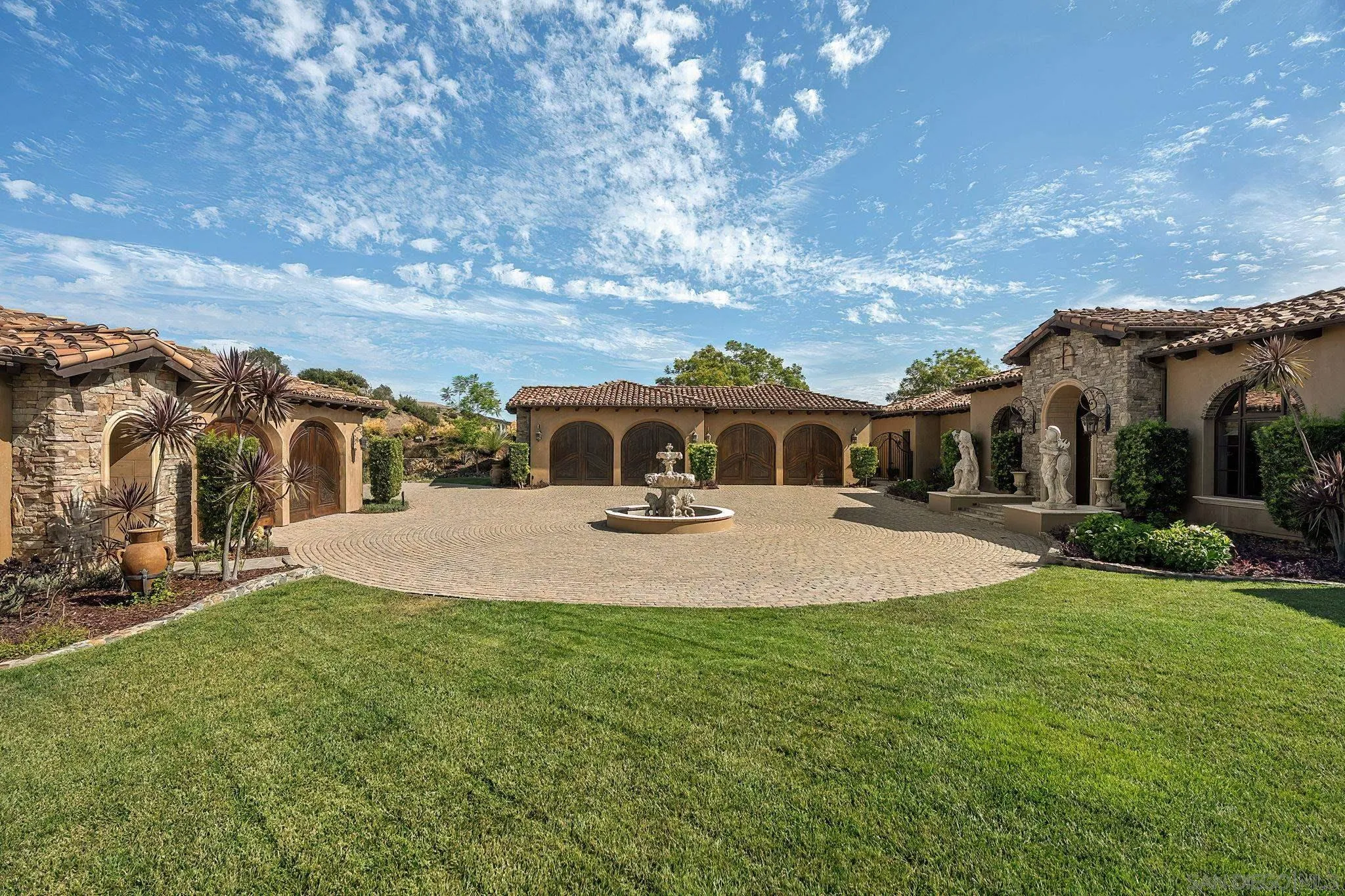 Mediterranean-style estate courtyard with arched garage doors, a central fountain, statues and a broad lawn.