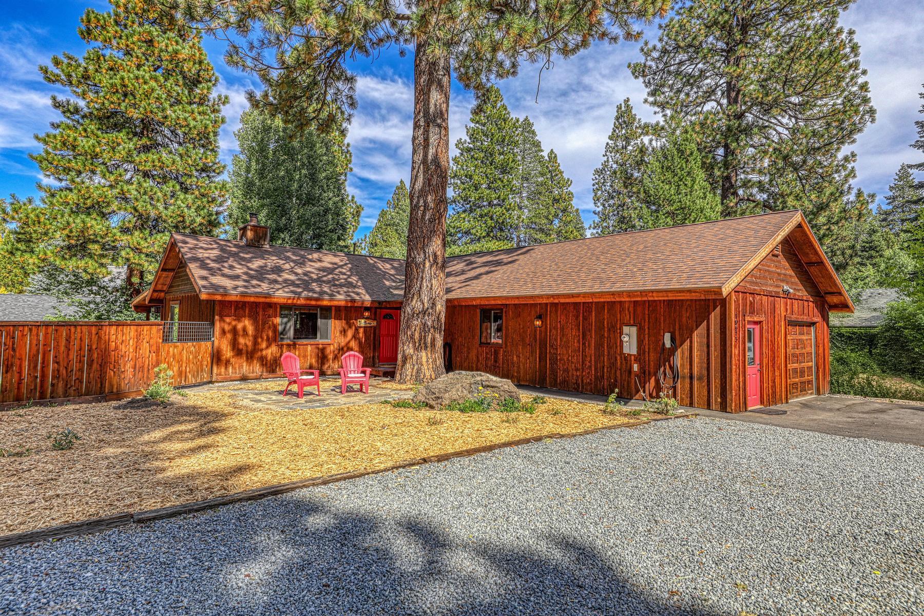 Wood-sided home among pine trees, with red doors, Adirondack chairs, and a gravel driveway.