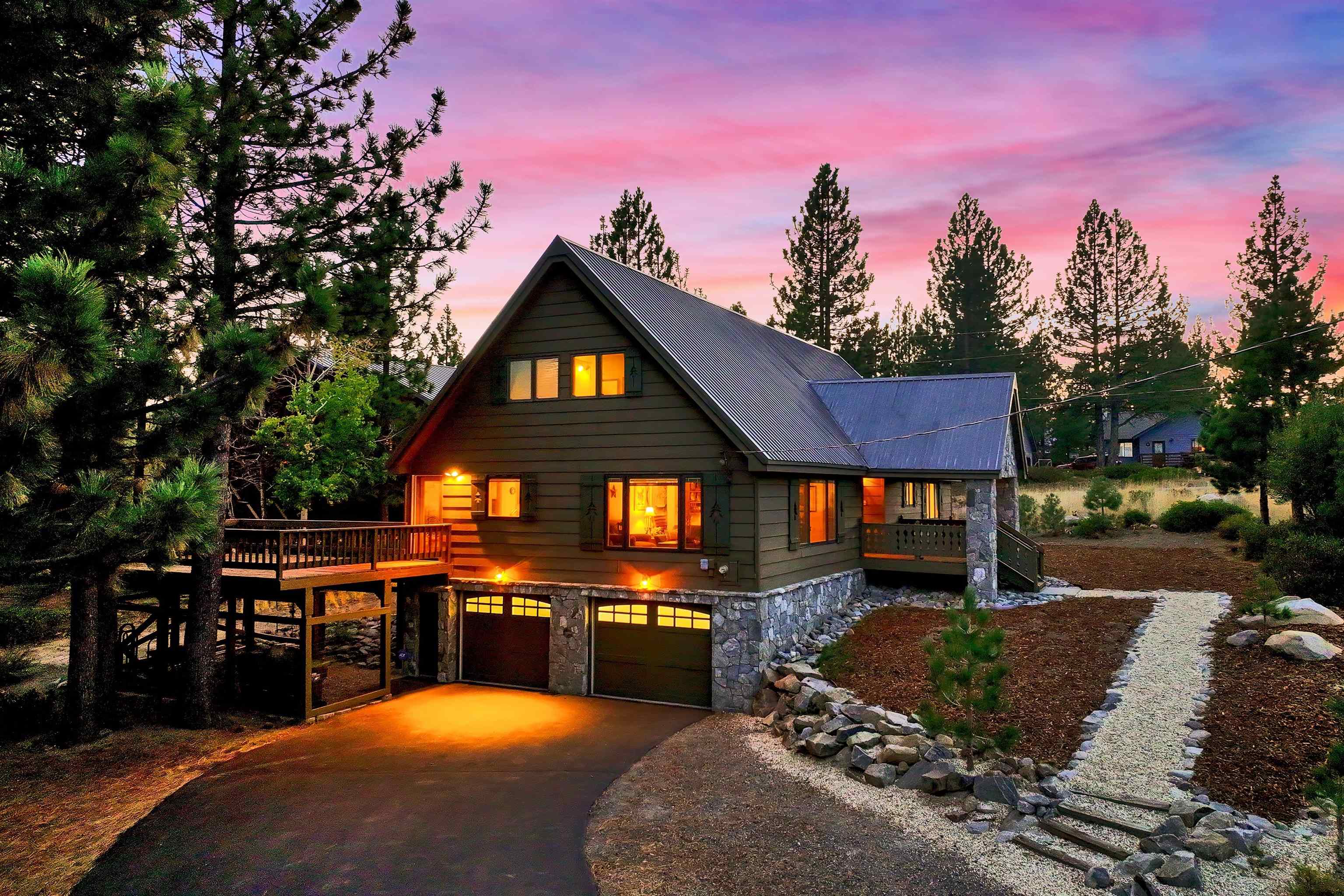 Mountain-style home with glowing windows and raised decks among pine trees at dusk.