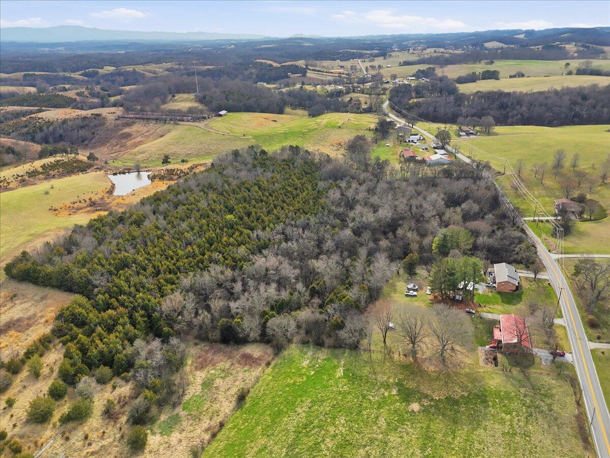 Aerial view of wooded acreage, open fields, a pond, and a road beside scattered buildings.