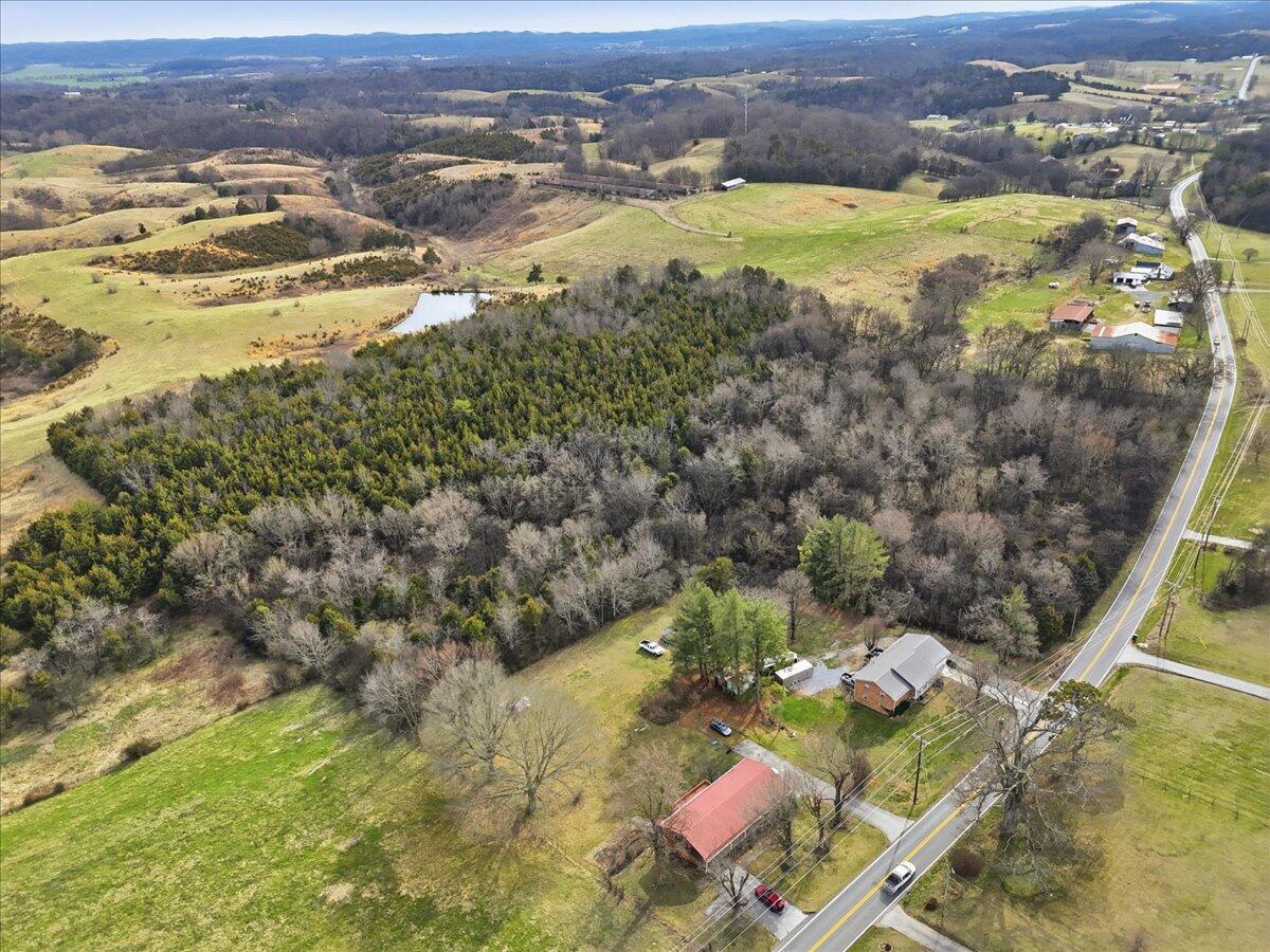 Aerial view of wooded acreage, open pasture, a pond, and buildings beside a rural road.