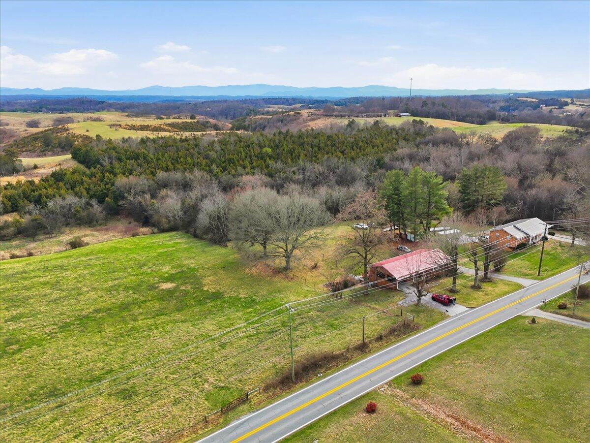 Aerial view of rural acreage with open fields, wooded hills, two roadside buildings, and distant mountain ridges.