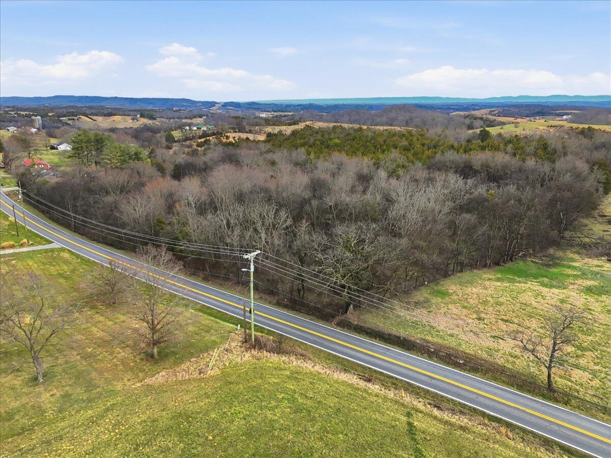 Aerial view of wooded rural land beside a road, with open fields and rolling hills beyond.