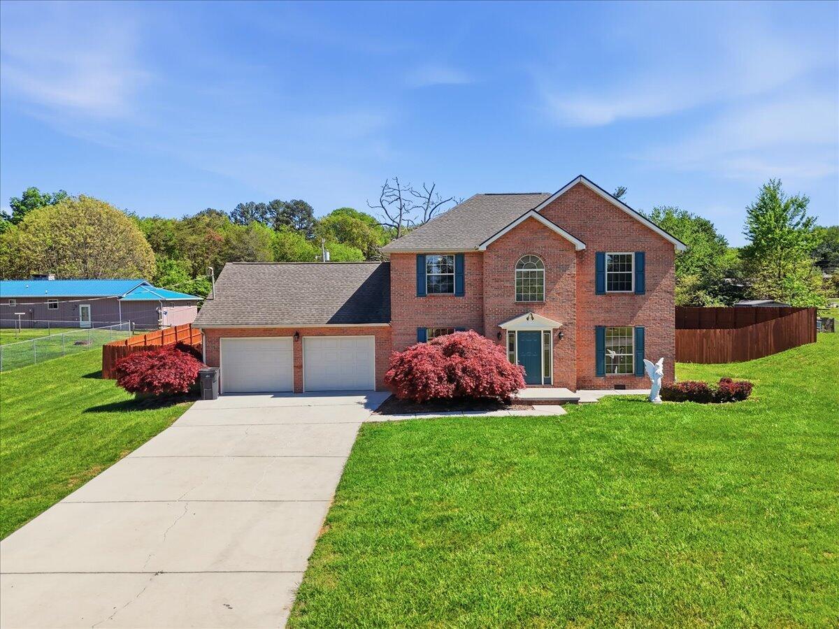 Two-story red brick house with an attached garage, broad driveway, and red ornamental shrubs.