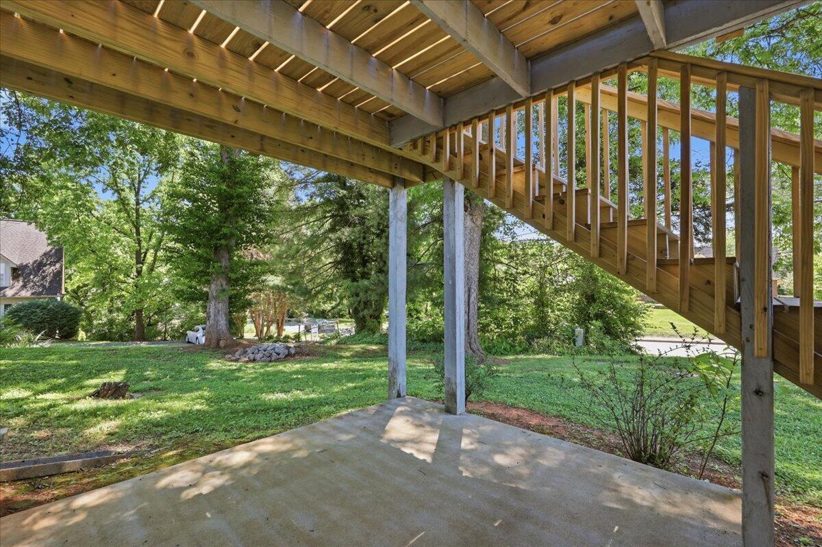Concrete patio beneath a raised wooden deck and long staircase, overlooking a leafy lawn.