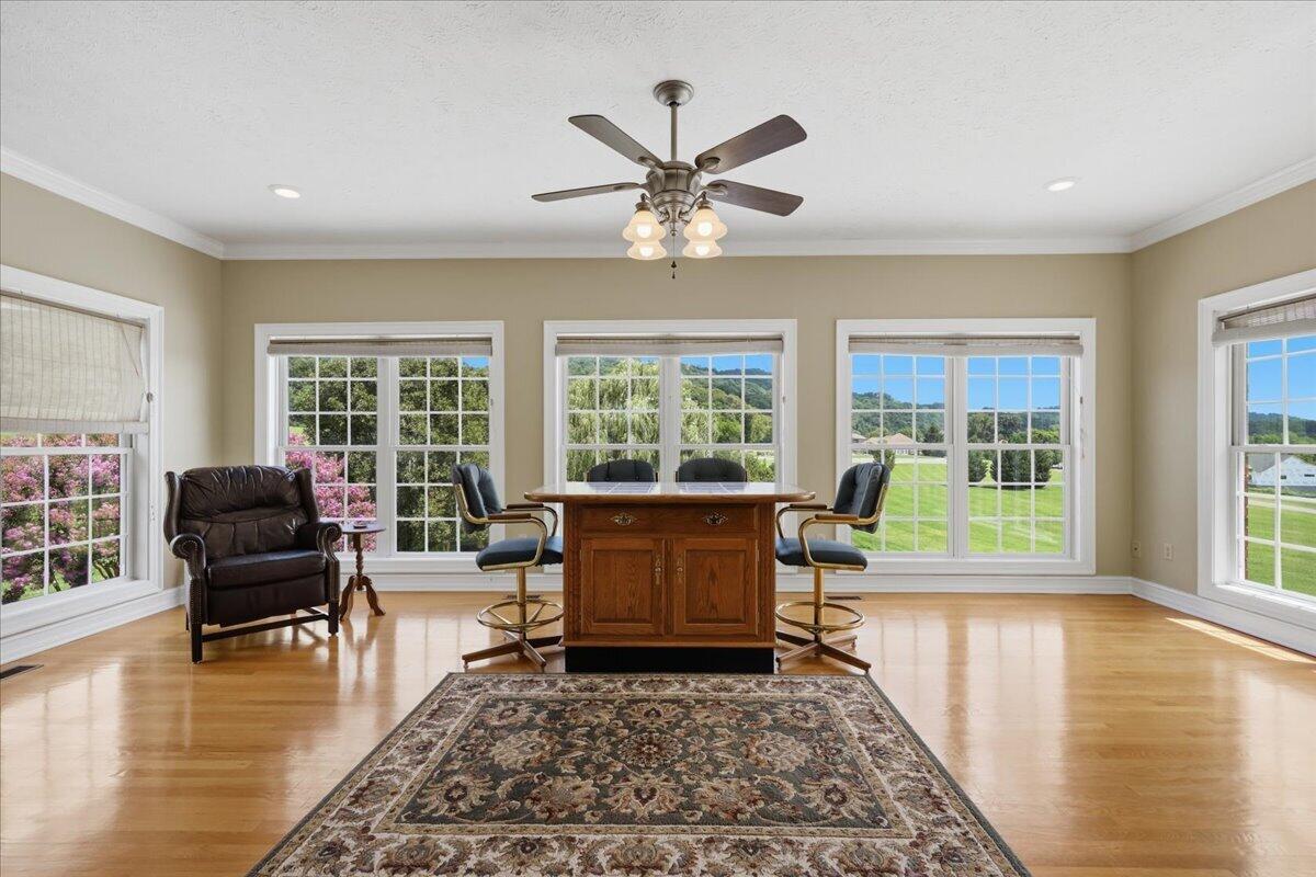 Furnished room with a wooden desk, office chairs, leather recliner, and large windows overlooking green hills.