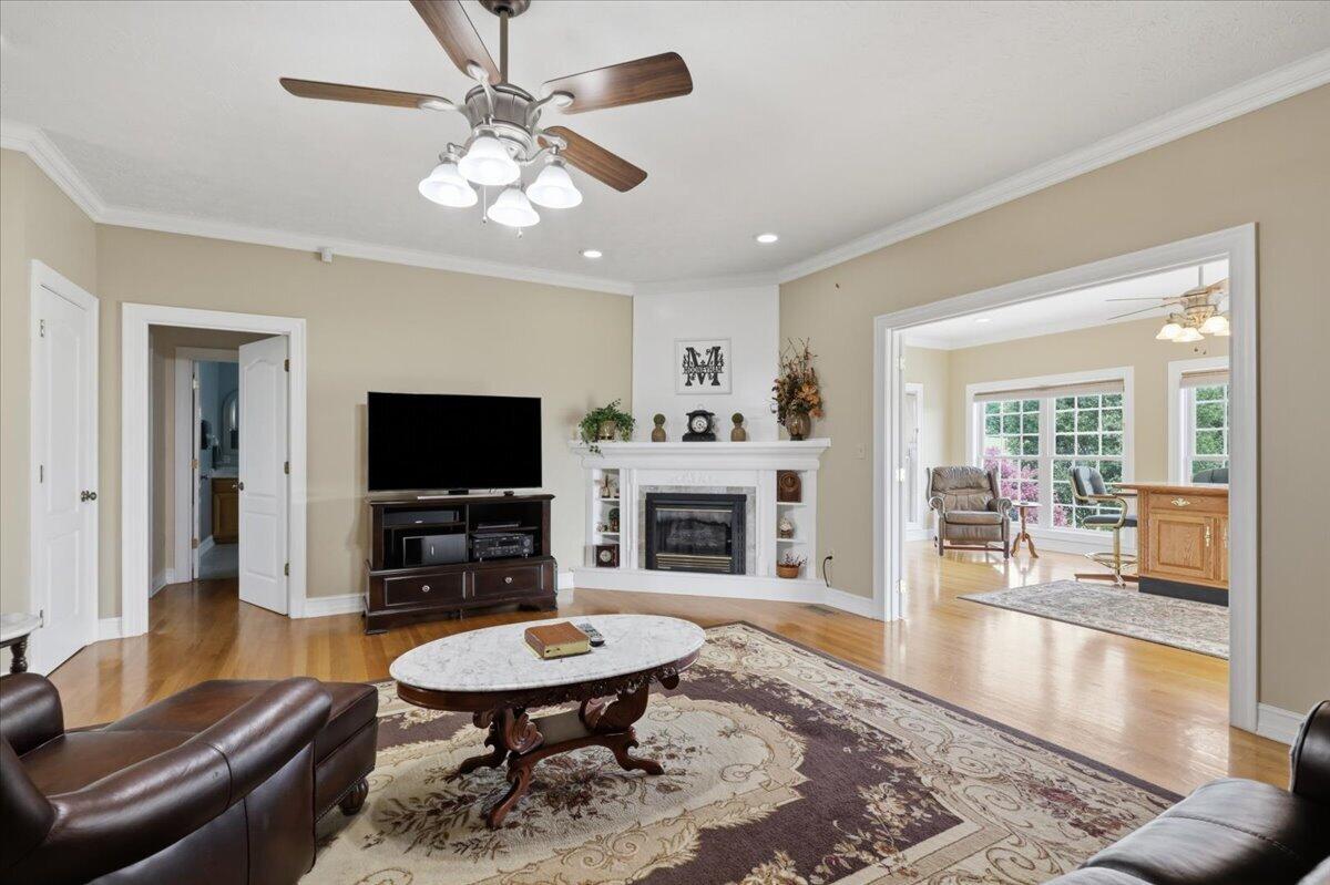 Living room with a white fireplace, ceiling fan, hardwood floors, and a wide opening to a bright adjoining room.