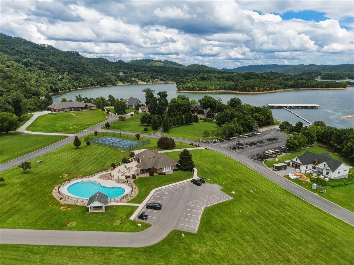 Aerial view of lakeside homes with a pool, tennis court, and dock beside wooded hills.