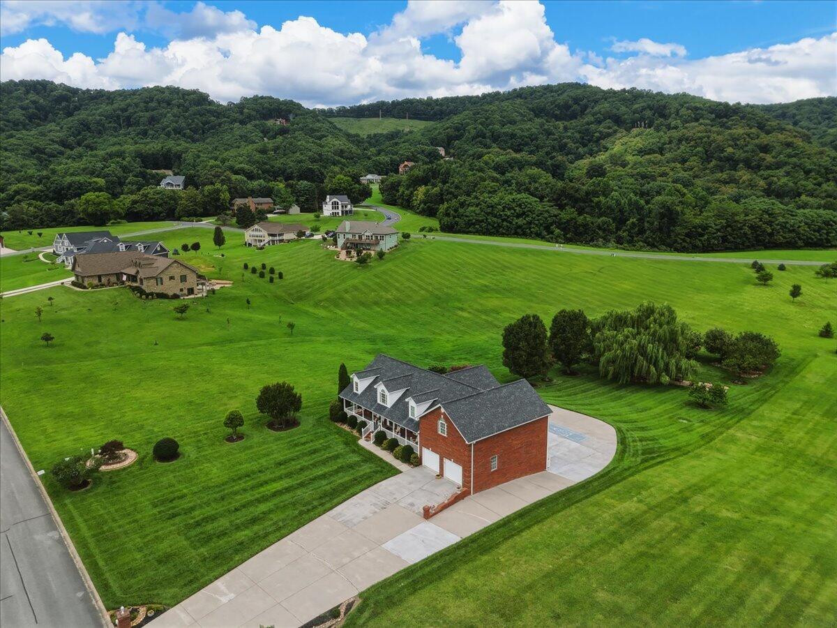 Brick house with a broad driveway and manicured lawn, viewed from above with wooded hills beyond.