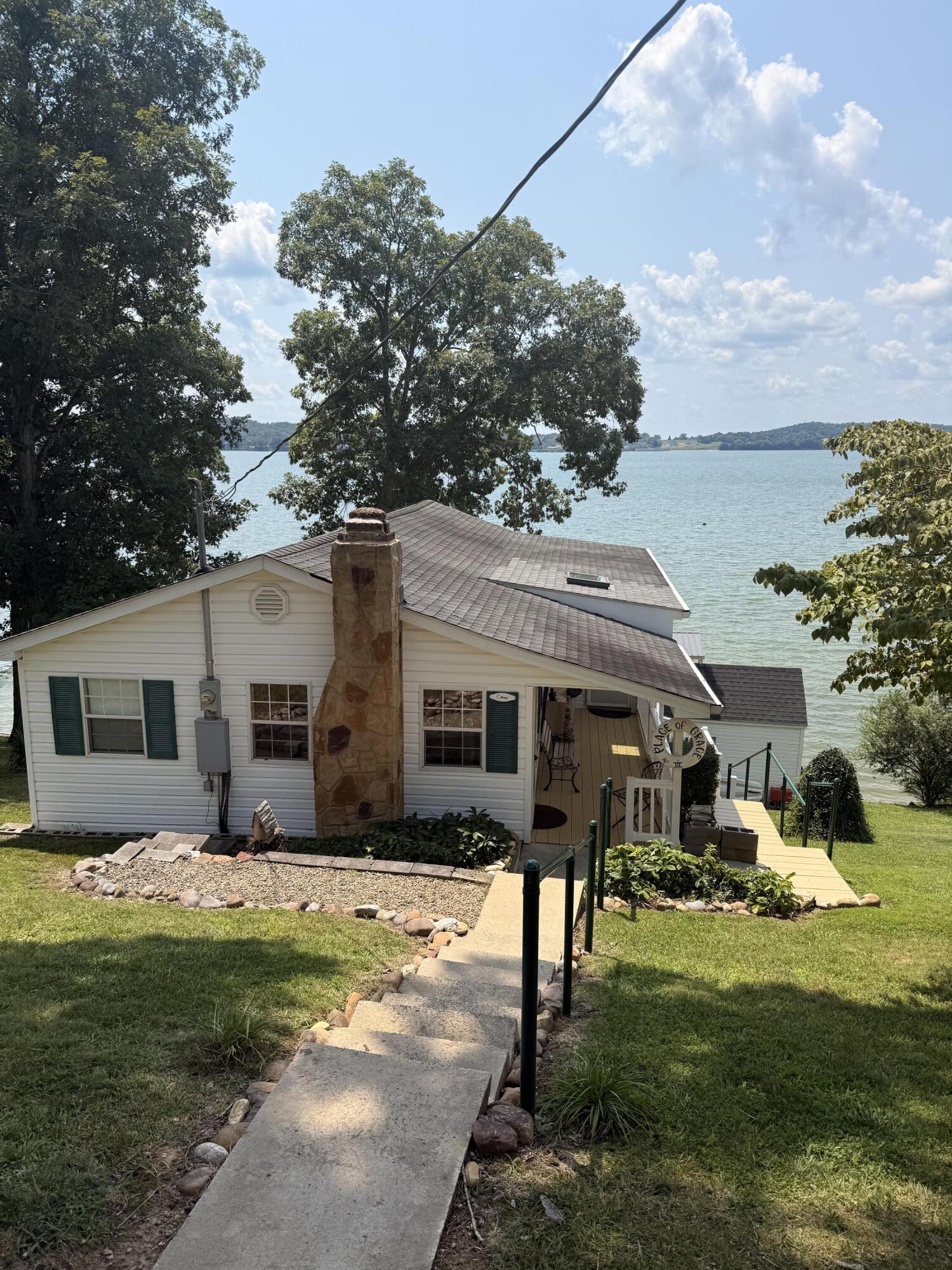 White lakeside cottage with green shutters, a stone chimney, and a path leading down toward the entrance.