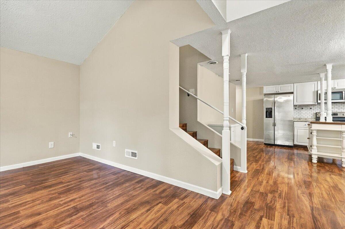 Open living area with hardwood floors, staircase, and adjoining kitchen with stainless steel appliances.