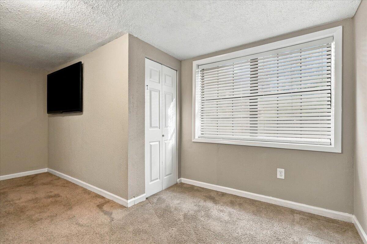 Mostly empty carpeted bedroom with a white closet door, wide window with blinds, and wall-mounted television.