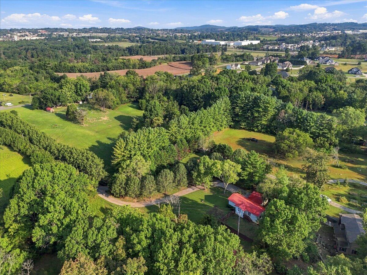 Aerial view of a wooded property with open lawns, a red-roofed outbuilding, and distant hills.