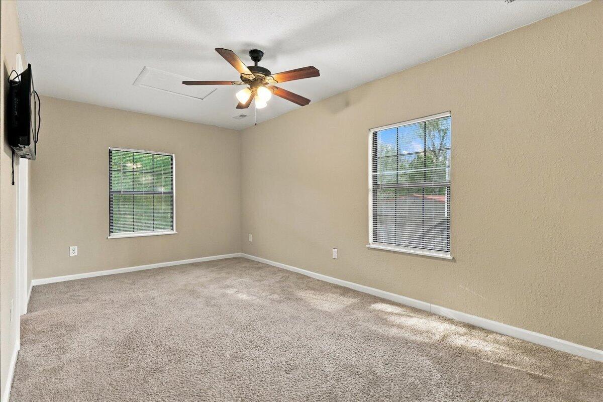 Empty carpeted room with beige walls, two windows, a ceiling fan, and a wall-mounted television.