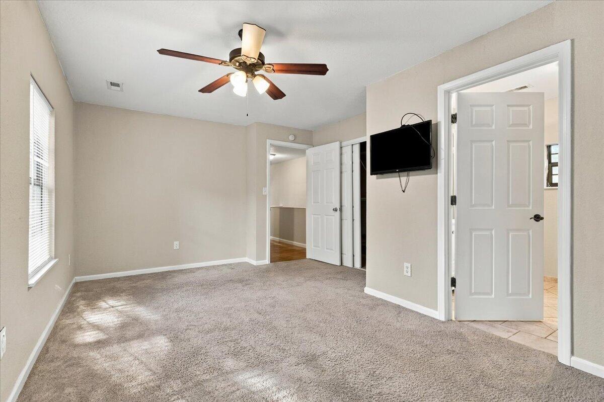 Empty carpeted bedroom with a ceiling fan, wall-mounted television, and sunlight through a window.