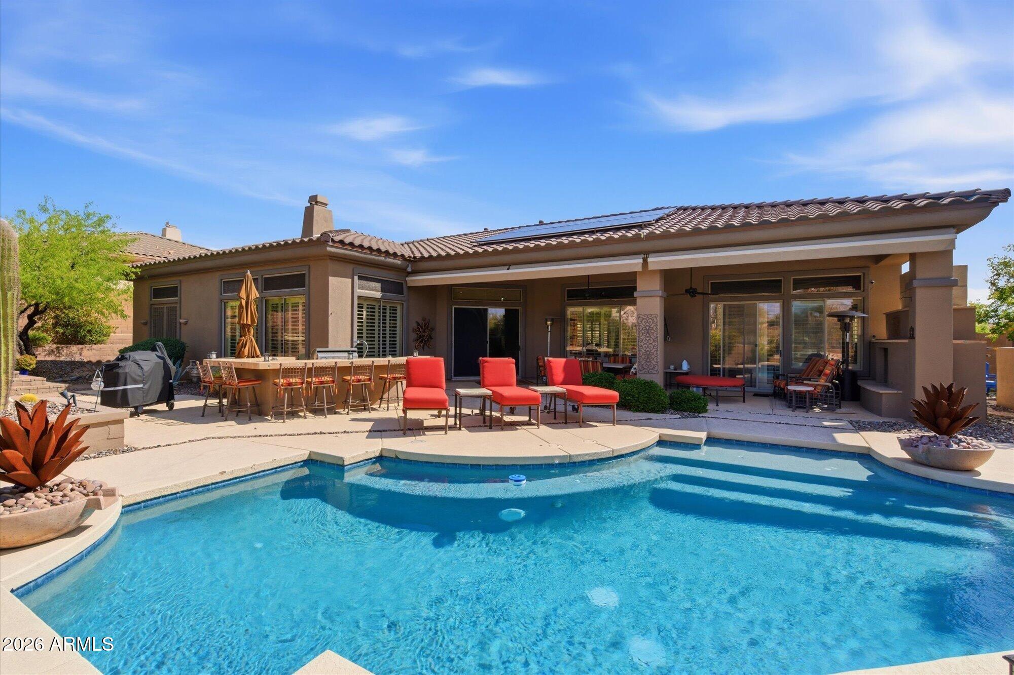 Backyard pool and furnished patio with red lounge chairs and solar panels on the roof.