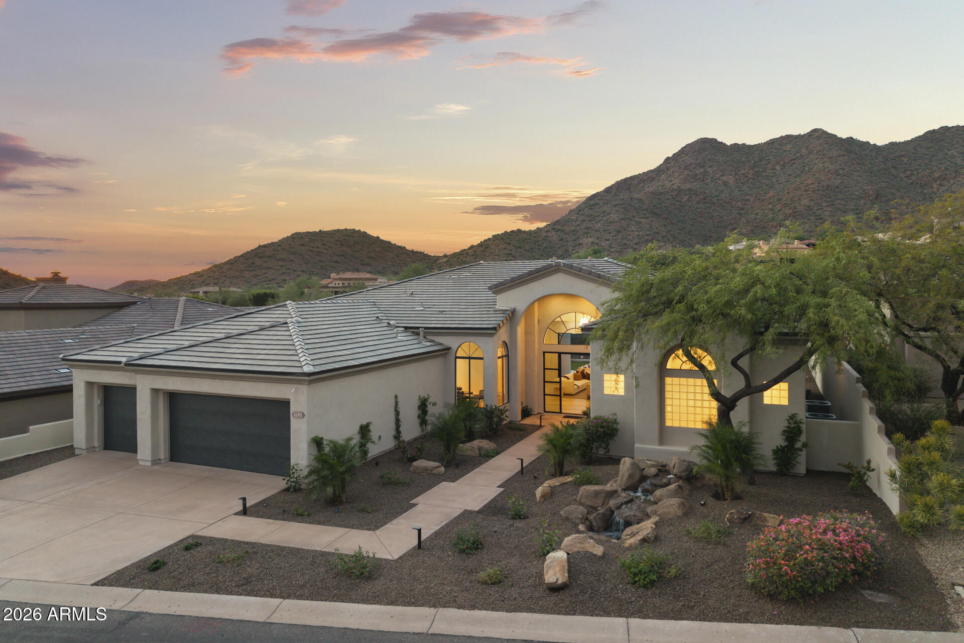 Stucco home with glowing arched windows, desert landscaping, and mountains at dusk.