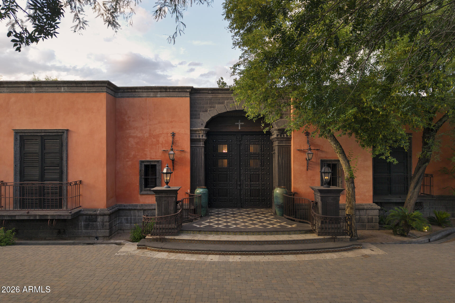 Terracotta stucco home with a grand arched stone entry, dark double doors, tiled steps, and mature trees.
