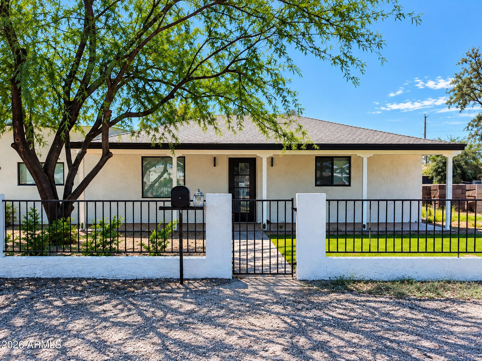 Single-story house behind a black metal fence, with a central gate and a large shade tree.