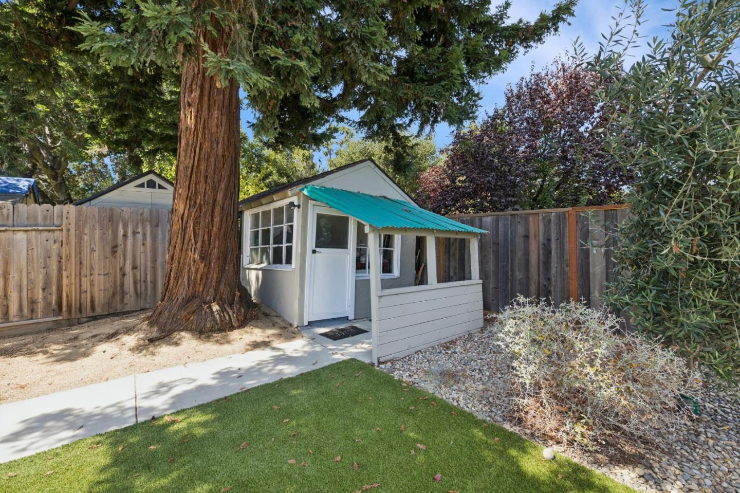 Backyard shed with a turquoise awning beside a large tree and fenced lawn.