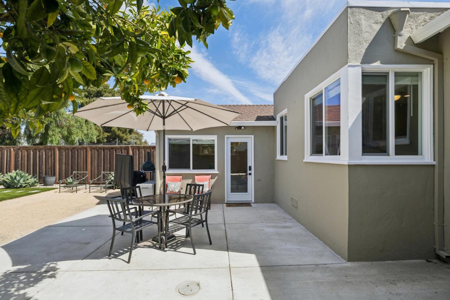 Backyard patio with outdoor dining set beneath a beige umbrella beside a gray stucco home.