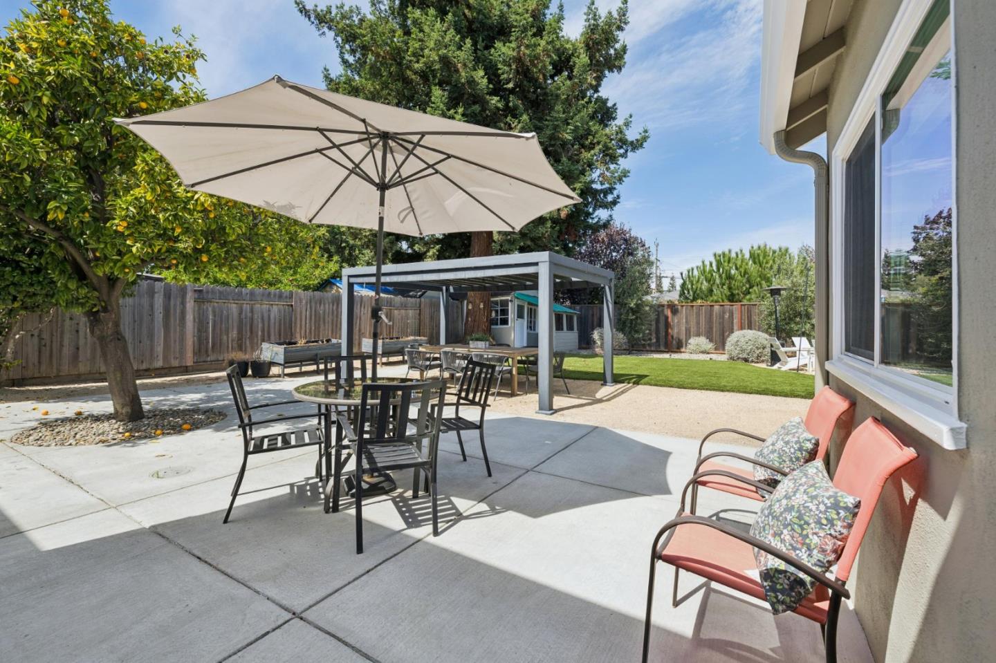 Backyard patio with umbrella-shaded dining table, pergola, cushioned chairs, and lawn.