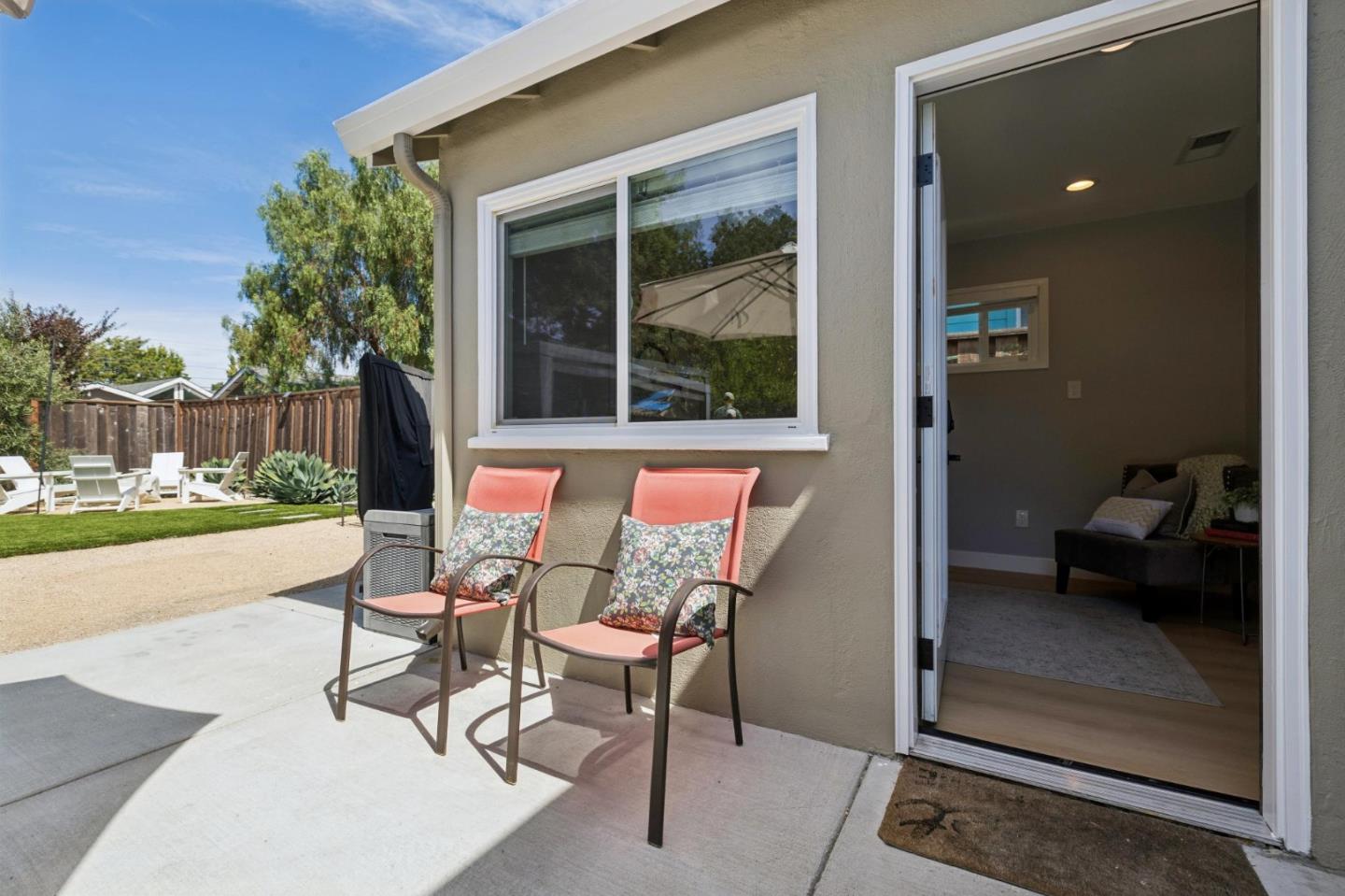 Patio with coral chairs beside an open doorway to a furnished room and fenced lawn.