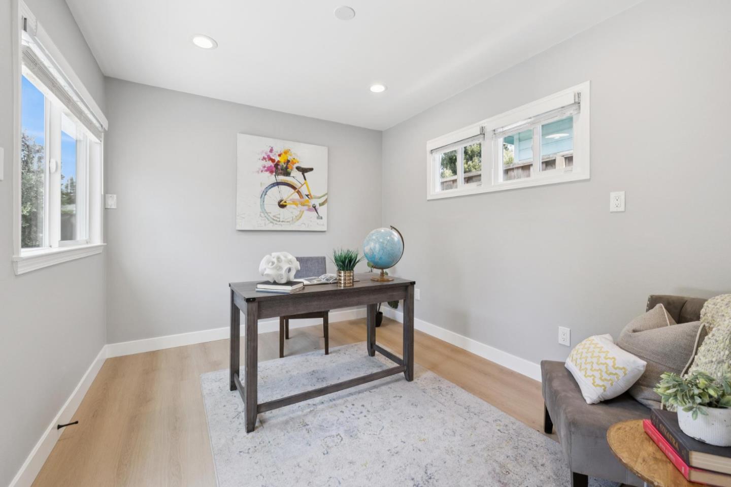 Furnished home office with a dark desk, windows on two walls, and colorful bicycle artwork.