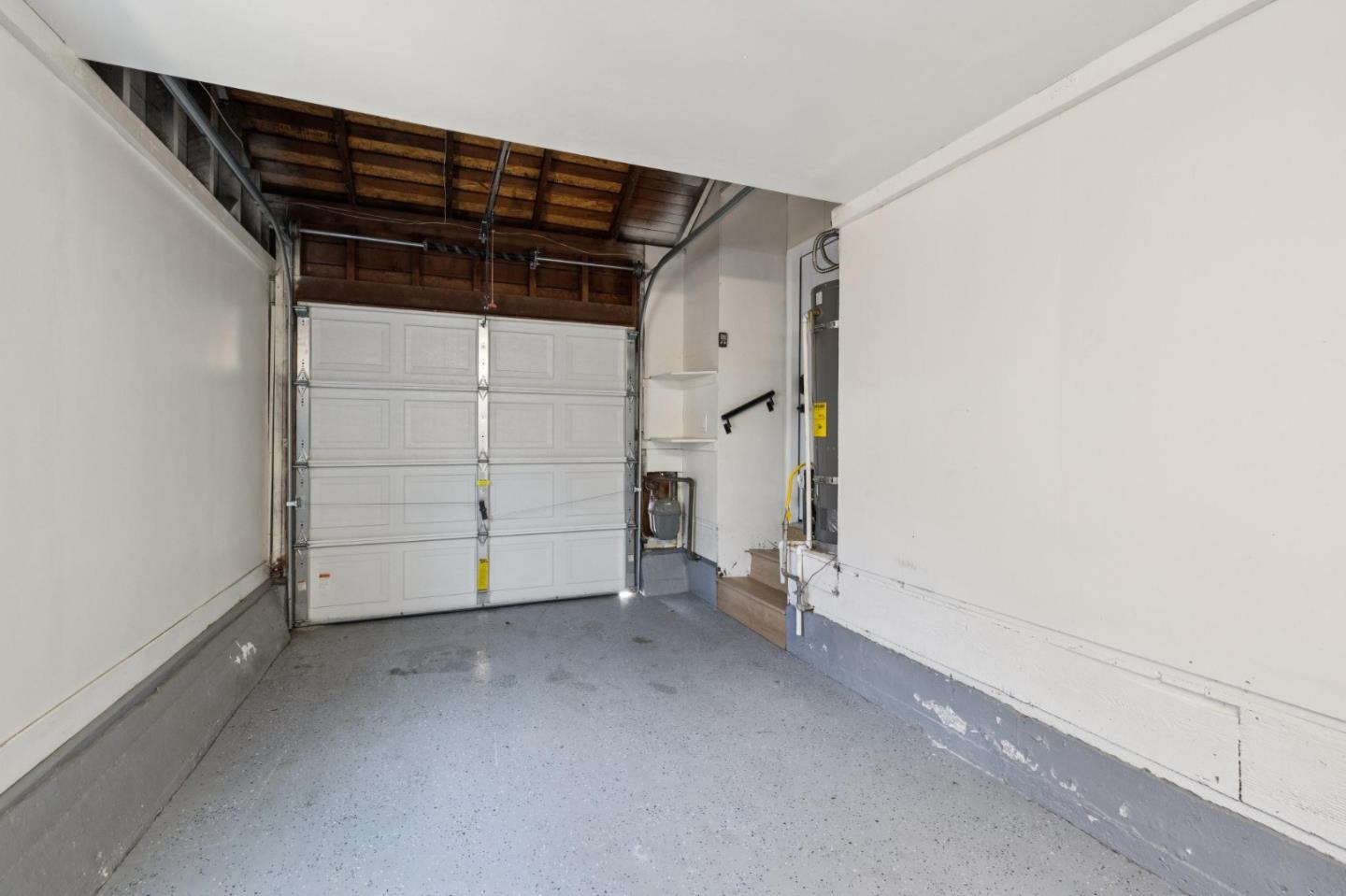 Empty garage with a closed sectional door, exposed wood rafters, and speckled gray floor.