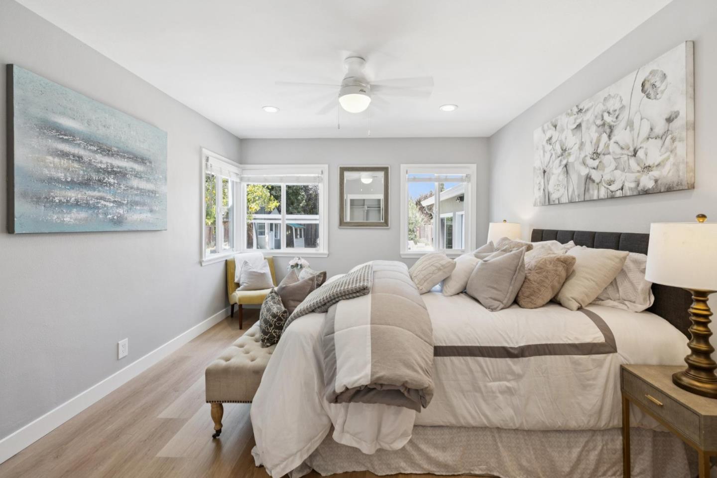 Staged bedroom with a large white bed, floral artwork, and windows overlooking greenery.