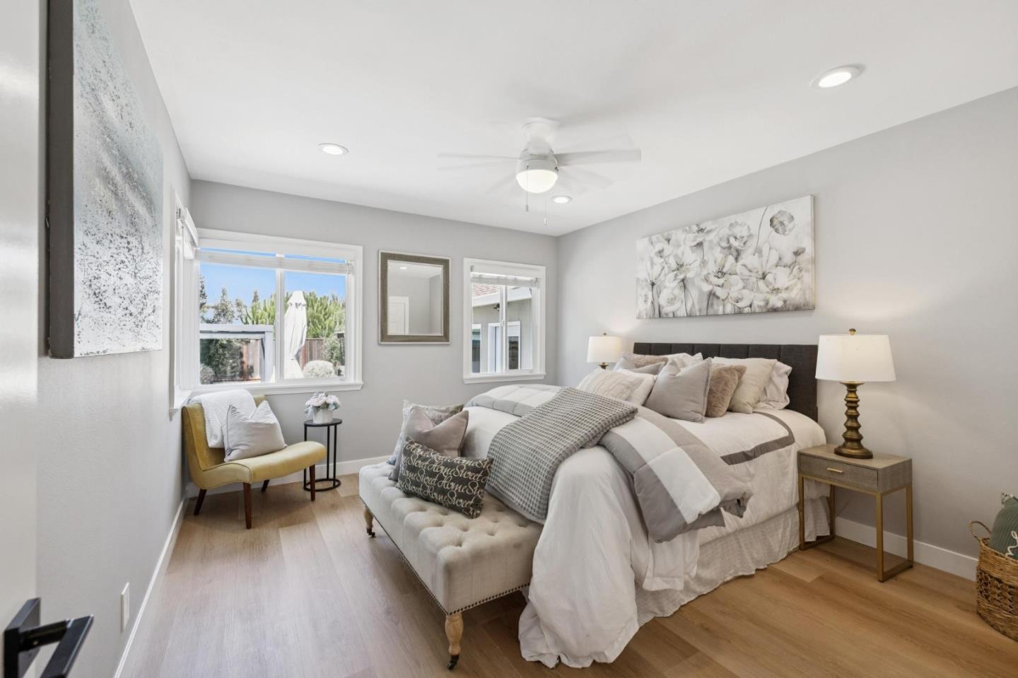 Staged bedroom with a large white-and-gray bed, tufted bench, mustard accent chair, and windows overlooking greenery.