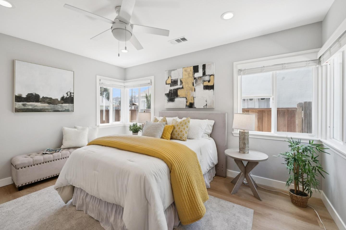 Staged bedroom with a white bed, mustard-yellow quilt, and large windows.