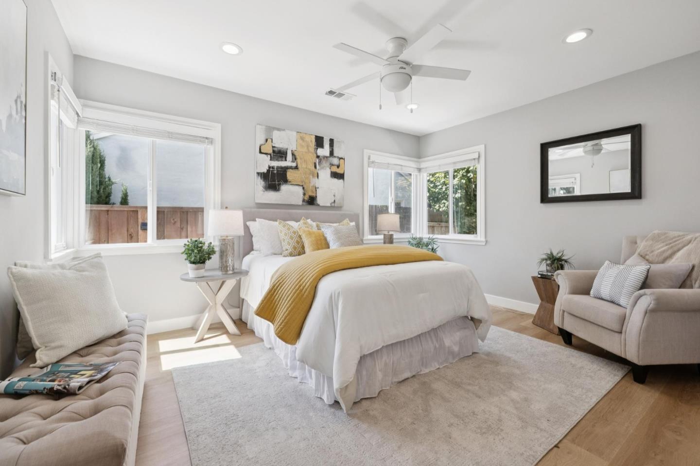 Staged bedroom with a white bed, mustard-yellow throw, ceiling fan, and large windows.
