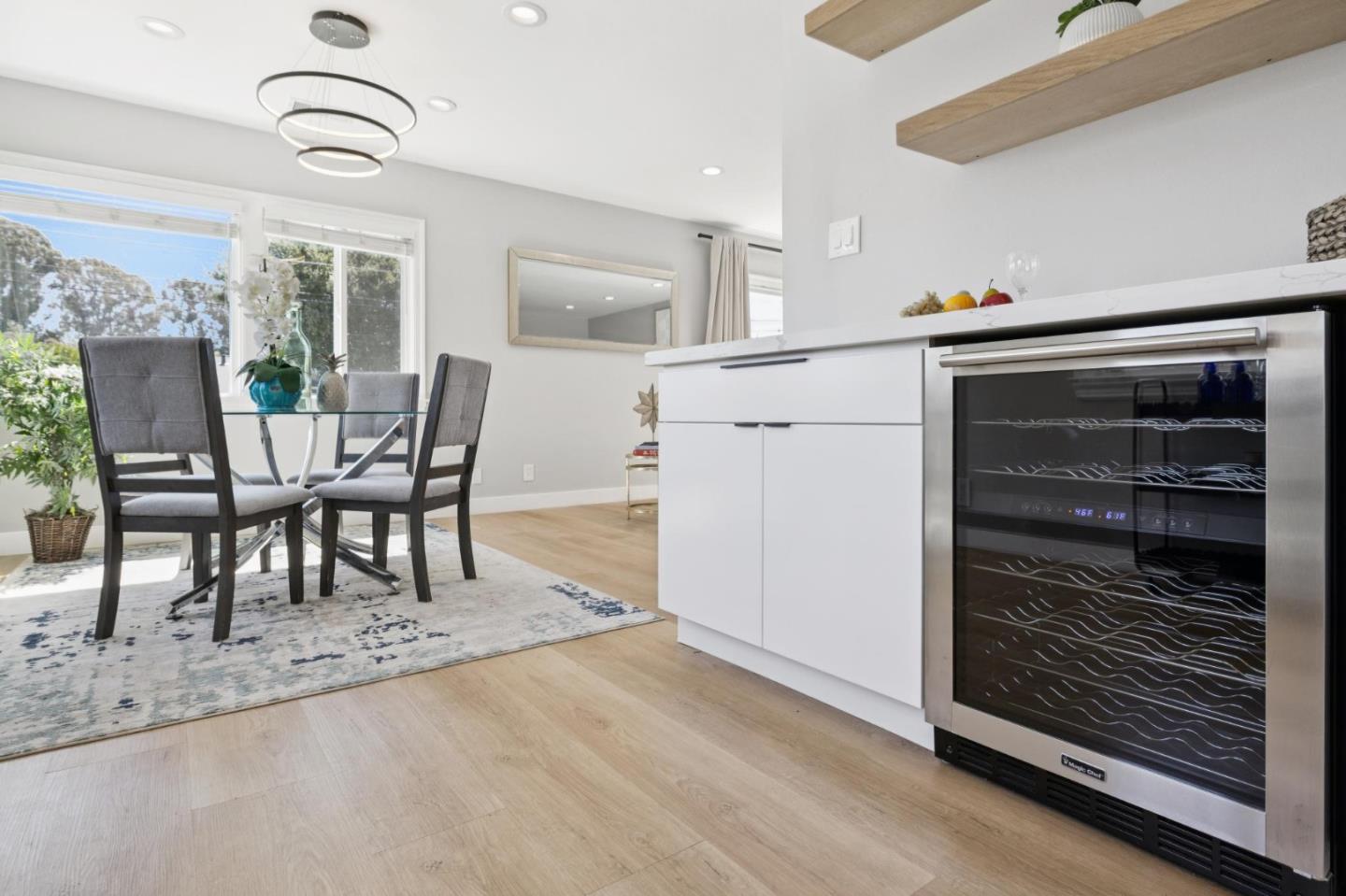 Kitchen and dining area with a white island, glass-front wine refrigerator, and window-side dining table.