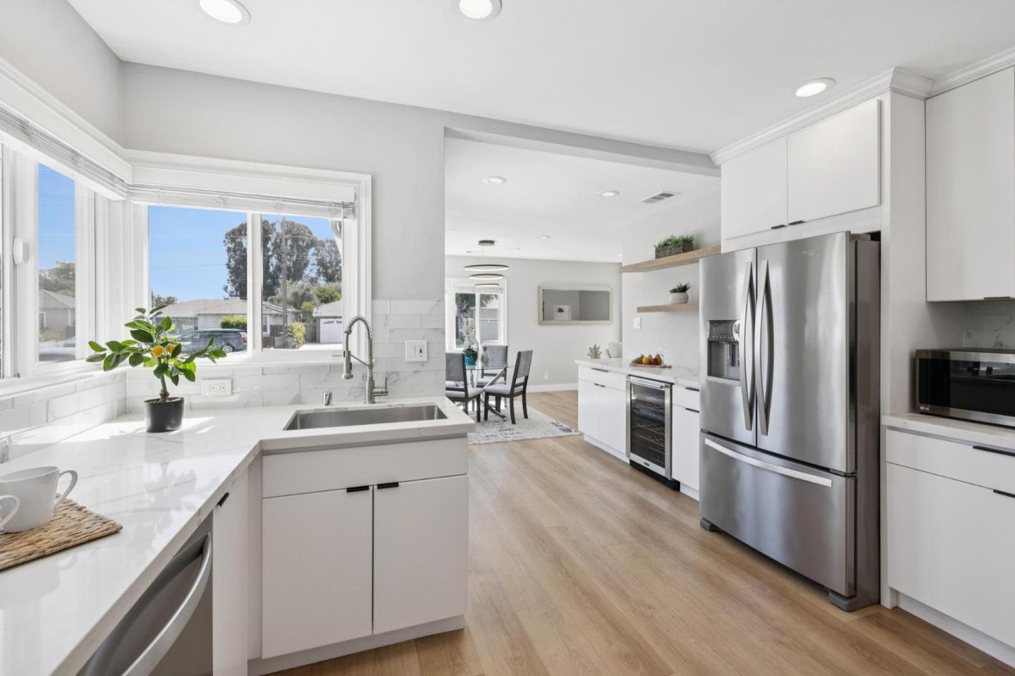 Bright white kitchen with a large window-side sink, stainless steel refrigerator, and light wood floors.