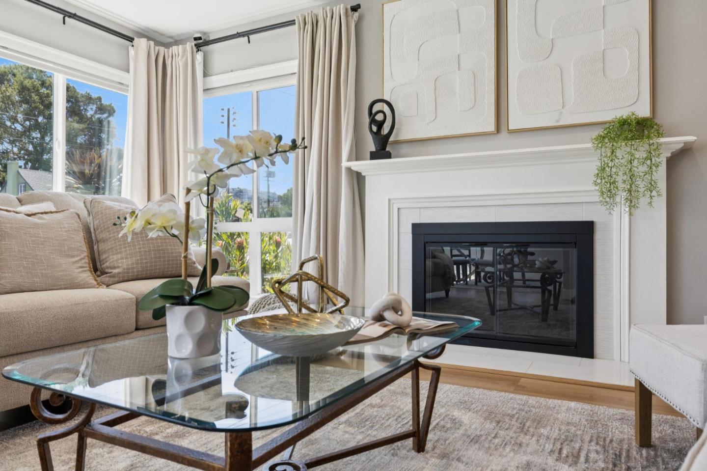 Living room with a white fireplace, beige sofa, and glass coffee table beside large curtained windows.