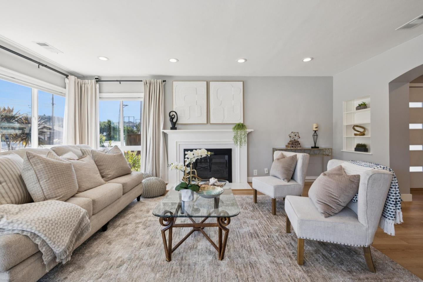 Staged living room with a white fireplace, large curtained windows, and neutral seating.