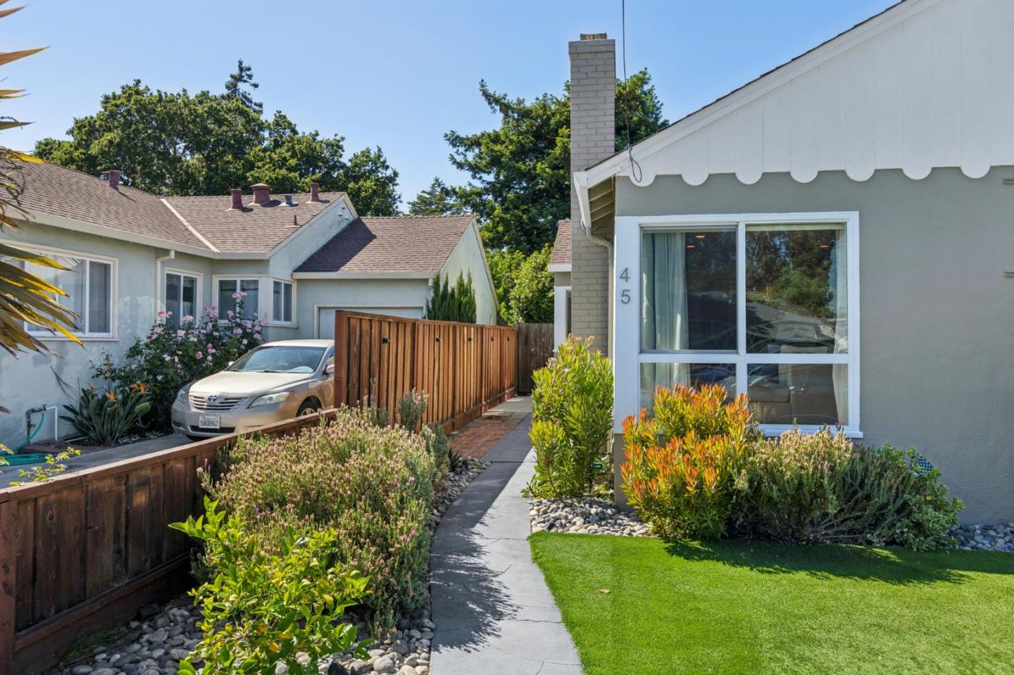 Home exterior with a large window, landscaped lawn, flowering shrubs, and a paved walkway.