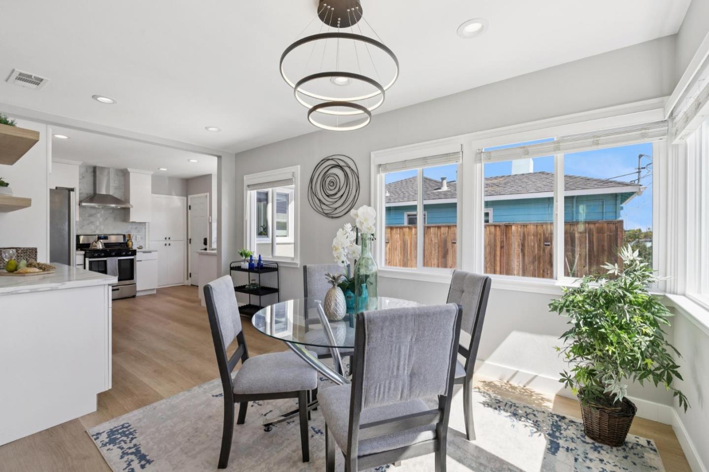 Dining area with a glass table and large windows, adjoining a white kitchen.